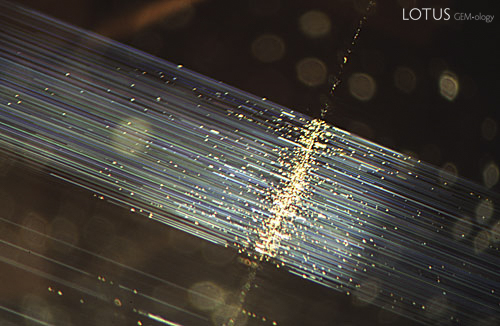 Figure 2. When fiber-optic illumination is used on the same chrysoberyl, bundles of fine light-scattering silky needles are seen extending at 90˚ from the linear growth band which is also more clearly detailed now. With fiber optics a tremendous amount of detail is gained. Magnified 10&times;. Photomicrograph &copy; John I. Koivula, microWorld of Gems.