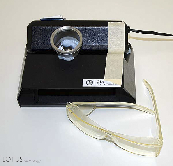 Figure 6. Setup for close-up examination of UV fluorescence. A small piece of blutac or clay inserted between the UV lamp and the viewing cabinet creates a small gap. This allows the stone to be viewed while positioned extremely close to the lamp, greatly increasing the ability to catch weak reactions. A lens is positioned to magnify the stone during viewing. The masking tape keeps the lamp from tipping off the cabinet. Special UV protection glasses such as those in front of the cabinet should be worn to protect the eyes from harmful SW radiation. Photo: Richard W. Hughes; Nikon D70